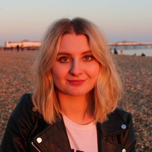 Headshot of artist Alex Cole, the founder of Lazy Lemon art studio. She is stood in the sun, on the beach with Brighton Pier blurred in the background.