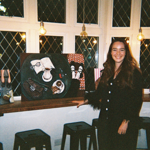 Megan Cleasby, female artist, stood next to two of her paintings propped up against a window of a pub.