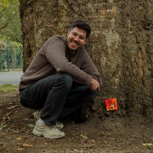 A picture of artist Tom Charter, kneeling by a large oak tree next too one of his small paintings that says "lucky sh*t" on it. 